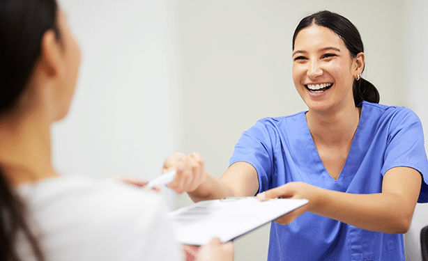 Smiling dental professional in blue scrubs handing paperwork to patient in clinical office setting.