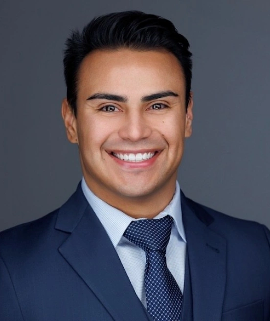 A portrait of a smiling man with short dark hair and brown eyes, wearing a navy blue suit jacket with a matching vest, a light blue checkered shirt, and a dark blue patterned tie against a plain gray background.