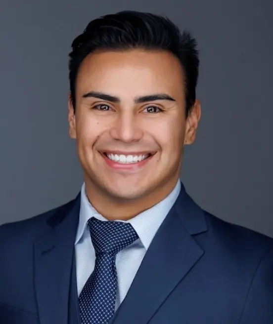 Professional portrait of a man with short dark hair and a wide smile. He is dressed in a dark blue suit, a light blue checkered button-down shirt, and a dark patterned necktie, set against a solid gray background.