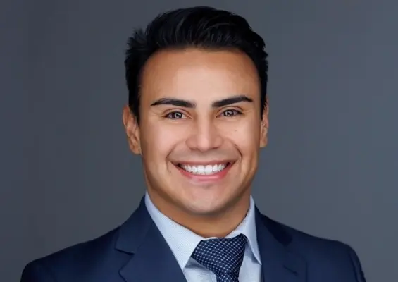 Professional headshot of a man with short dark hair, smiling warmly at the camera. He is wearing a dark blue suit jacket, a light blue checkered dress shirt, and a dark patterned tie against a smooth gray studio background.